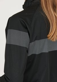 Schwarze Jacke mit einem strukturierten Design und einem horizontalen grauen gepunkteten Streifen im Bereich des Oberarms. Hoher Kragen und glattes Material.