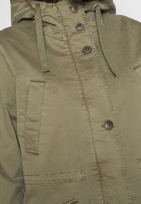 Oliwkowa, kapturowa kurtka o gładkiej fakturze, z guzikami, sznurkiem ściągającym oraz dwiema przednimi kieszeniami. Wykonana z trwałej tkaniny.