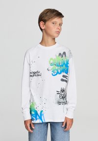 Biała koszulka z długim rękawem z kolorowymi wzorami graffiti w kolorach zielonym, niebieskim i czarnym, z efektami splash. W połączeniu z jasnoniebieskimi dżinsami.