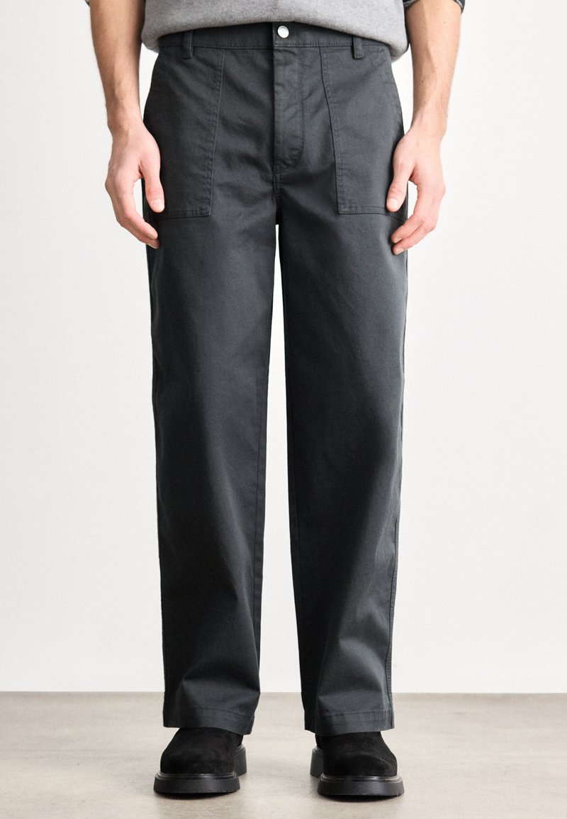 Sötétszürke, egyenes szárú nadrág elülső zsebekkel, gombos záródással és sima tapintású anyagból. Fekete lábbelivel viselve.