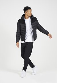 Chaqueta negra acolchada con un acento texturizado negro en el hombro, combinada con una camiseta blanca y pantalones negros. Modelo lleva zapatillas blancas.