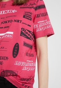 Červená košile s černým a bílým textem na téma cestování a letiště, včetně názvů měst jako Tokio, Glasgow, Praha a Moskva.