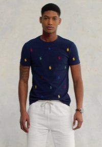 Joven vestido con una camiseta azul marino con pequeños logotipos multicolores y pantalones blancos con cordón, de pie frente a una pared clara.