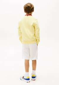 Sudadera amarilla con cuello rojo, shorts blancos y zapatillas blancas con detalles en azul y amarillo. Vista desde atrás.