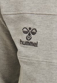 Tessuto grigio della felpa con una superficie testurizzata, caratterizzato da un logo ricamato nero con design simmetrico e il nome del marchio "hummel."