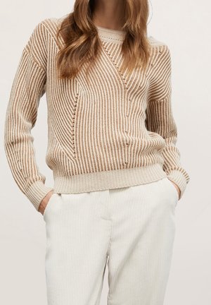 Mujer con suéter de punto beige y crema con rayas y pantalones texturizados blanco roto, con las manos en los bolsillos, sobre un fondo liso.