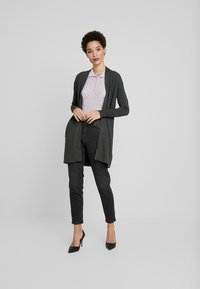 Grafitgrauer Cardigan mit weicher Textur, offenes Design, kombiniert mit einem helllila strukturierten Hemd, schwarzen Jeans und schwarzen High Heels.