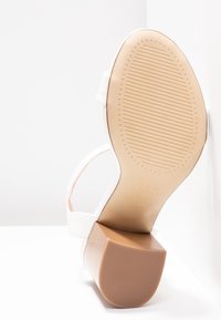 Sandale à talon haut blanche avec une semelle extérieure beige texturée, des brides étroites et un talon bloc. Le design présente des détails minimalistes.