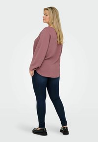 Rožinė ilgomis rankovėmis palaidinė su apvaliu apačiu, derinama su tamsiai mėlynomis liesomis džinsomis. Modelis vilki juodus batus, demonstruodamas laisvą siluetą ir švelnią tekstūrą.