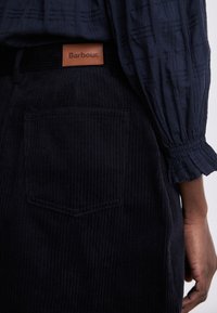 Dunkle Cordhosen mit vertikaler Rippung, versehen mit einem braunen Lederetikett mit eingraviertem "Barbour" am Taillenbund und einer einzelnen Gesäßtasche.