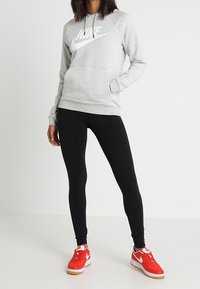 Šedá mikina Nike s bílým logem, kombinovaná s černými legínami a červenými teniskami. Má kapuci a boční kapsy.