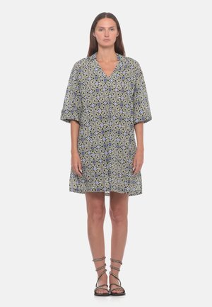 Donna con i capelli lunghi indossa un vestito ampio al ginocchio con un motivo geometrico dettagliato e sandali neri piatti con cinturini, in piedi su uno sfondo semplice.
