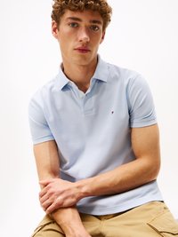 Világoskék pólóing rövid ujjakkal, gallérral és egy kis logóval a bal mellrészen. A modellen középhosszú, göndör haj van.