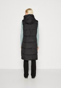 Zwarte gewatteerde vest met capuchon, voorzien van verticale naden en een lange snit. Gelaagd over een lichtblauw sweater en zwarte broek.