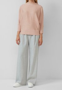 Femme portant un pull ample rose clair, un pantalon large gris clair et des baskets blanches, debout devant un fond uni.
