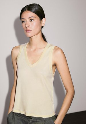 A young woman poses in a light yellow sleeveless top, with long dark hair slicked back, against a plain wall, hands in pockets.