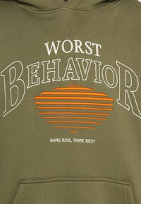 Felpa verde oliva con testo ricamato bianco e arancione: "WORST BEHAVIOR" sopra un grafico del sole, con "SOME RISE, SOME REST" sotto.