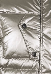 Section d'une veste matelassée argentée métallique avec un rembourrage matelassé, une poche à rabat avec un bouton noir, et des détails de couture visibles.