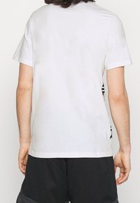 Bílá krátká trička s krátkým rukávem vyrobená z bavlny, s jednoduchou zadní stranou a grafickým designem na boku. Měkká textura, běžný střih.