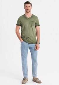 Olivově zelené tričko s véčkovým výstřihem, krátkými rukávy a přední kapsou, v kombinaci se světle modrými slim-fit džíny a béžovými mokasíny.