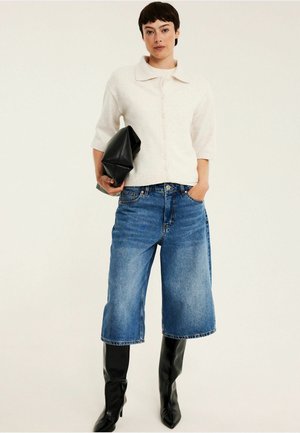 Femme portant un pull blanc boutonné, un short en jean large mi-genoux, des bottes noires montantes, tenant une pochette noire.