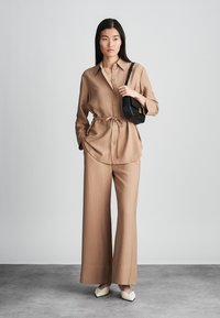 Camicia beige con cintura, maniche lunghe e pantaloni a gamba larga con righe verticali. Il modello tiene una piccola borsa nera con dettagli dorati.
