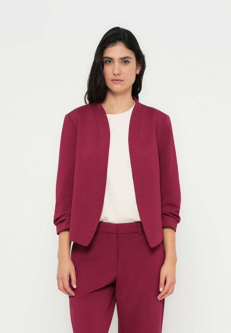 Σακάκι βουργουνδί με άνετη γραμμή, χωρίς γιακά και με φουσκωμένα μανίκια, συνδυασμένο με ανοιχτό μπεζ τοπ και παντελόνι στον ίδιο τόνο.