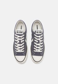 Ténis de lona azul-marinho com biqueiras e atacadores brancos. Possui um design clássico de atacadores e a marca "Converse All Star" na língua.