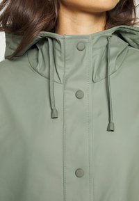 Olivgrüne wasserdichte Jacke mit Kapuze, ausgestattet mit Druckknöpfen, verstellbaren Kordeln und einer glatten, matten Textur.