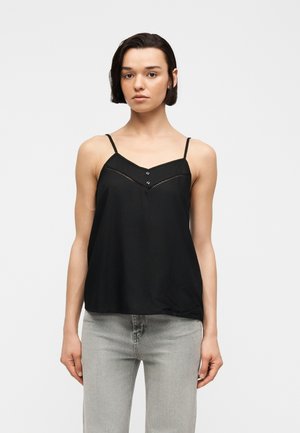 Kvinde med kort mørkt hår iført en sort ærmeløs top med tynde stropper og lysegrå jeans, stående mod en ensfarvet hvid baggrund.