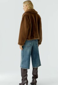 Vrouw in een korte bruine nepbontjas, met een wijde denim culotte en kniehoge donkerbruine leren laarzen, staande in zijaanzicht.