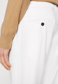 Pantaloni bianchi con una sola tasca posteriore dotata di chiusura a bottone nero. Mano appoggiata vicino alla tasca, con una manica di un maglione color beige visibile.