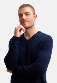 Mörkblå V-ringad tröja i mjukt material; har långa ärmar och ribbade manschetter. Modellen står i en eftertänksam pose.