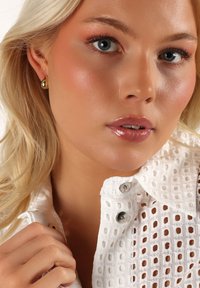 Vit broderad blus med silverknappar, en närbild av ett ansikte med naturlig sminkning, ljust blont hår och guldörhängen.
