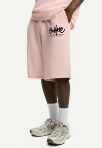 Rosa bomullsshorts med sidofickor, prydda med en svart grafisk tryck på vänster ben. I kombination med vita strumpor och ljusa sneakers.