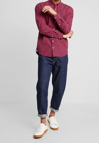 Camicia a maniche lunghe a quadri rossi e blu, abbottonata, abbinata a pantaloni in denim scuro con risvolto e sneakers bianche con suole marroni.
