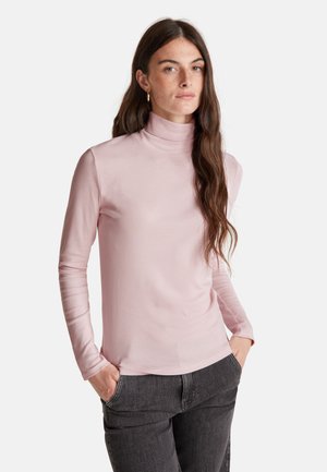 Donna con lunghi capelli castani che indossa orecchini a cerchio dorati, un maglione rosa chiaro a collo alto e jeans grigio scuro, con le mani in tasca.