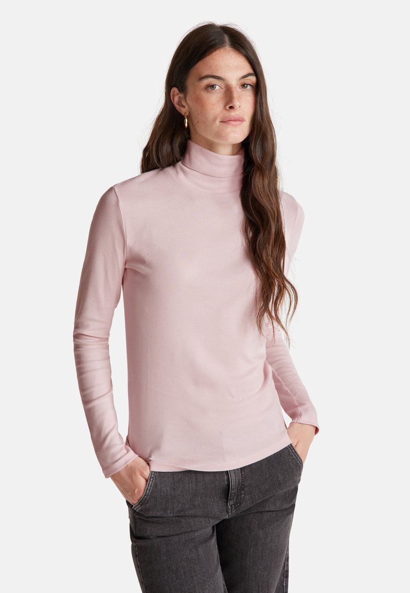 Frau mit langen braunen Haaren, die goldene Ohrringe, einen hellrosa Rollkragensweater und dunkelgraue Jeans trägt, mit Händen in den Taschen.