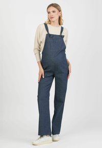 Ogrodniczki w ciemnoniebieskim kolorze, z regulowanymi ramiączkami, kieszenią z przodu i szerokimi nogawkami. W połączeniu z jasnobeżową bluzką z prążkowanej tkaniny i białymi sneakersami.