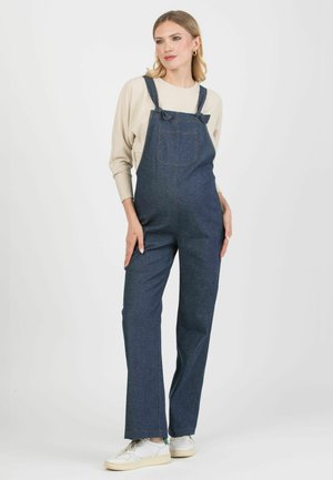 Dunkelblaue Jeanslatzhosen mit verstellbaren Trägern, einer Vordertasche und weitem Bein. Kombiniert mit einem hellbeigen, gerippten Oberteil und weißen Turnschuhen.