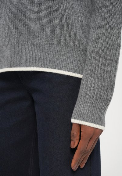 Pull en tricot gris avec une texture côtelée, présentant des finitions blanches aux poignets et à l'ourlet. Assorti à un pantalon en denim foncé.