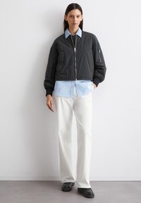 Schwarze Bomberjacke mit Reißverschluss, gerippten Bündchen und zwei Fronttaschen, über einem hellblauen gestreiften Hemd und weißen Hosen getragen. Schwarze Schuhe.