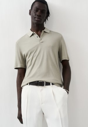 Jeune homme portant un polo beige à manches courtes rentré dans un pantalon blanc avec une ceinture noire, debout contre un mur clair uni.