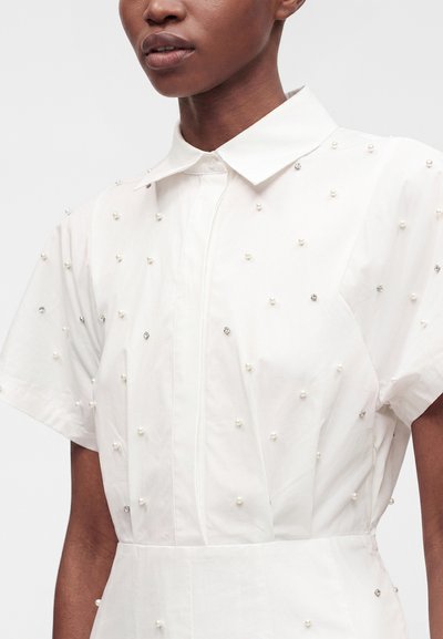 Gros plan d'une personne portant une chemise blanche à manches courtes avec col, décorée de petites perles et strass.