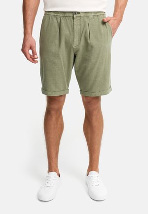 Man draagt olijfgroene knielange korte broek met opgerolde zomen, voorste knoop en ritssluiting, zijzakken en witte sneakers.