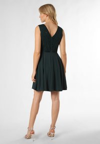 Dark green dress with lace bodice, V-back, sleeveless design, flared skirt, and a satin waistband. Worn with strappy high-heeled sandals.