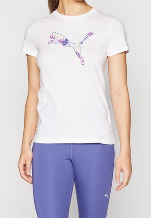 Kobieta w białej koszulce z kwiatowym wzorem logo Puma i fioletowych legginsach z małym białym logo Puma na udzie.