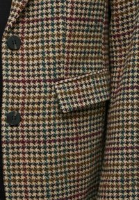 Checkered blazer in warm tones, featuring a houndstooth pattern with black buttons and a folded pocket detail. Textured wool fabric.