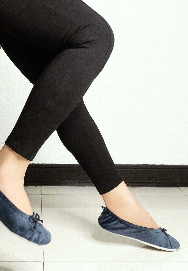 Ballerines en velours bleu marine avec un bout arrondi et un petit nœud, associées à des leggings noirs sur un sol carrelé.