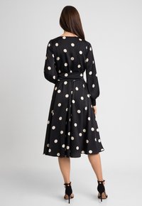 Czarna sukienka w kropki z długimi rękawami, dopasowaną talią i zwiewnym dołem. Wykonana z gładkiego materiału z delikatnym połyskiem.